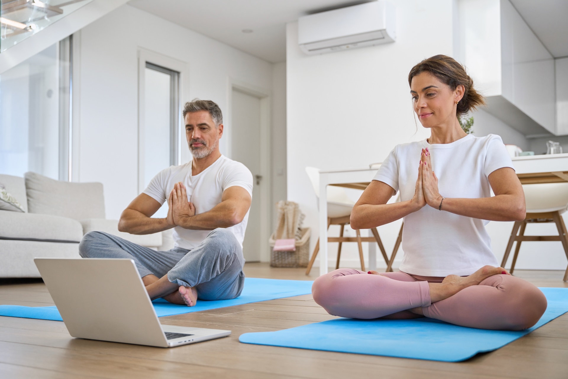 Middle aged family couple doing yoga at home looking at laptop. Healthy calm serene mature older man and woman doing exercises meditating watching wellness tutorial in living room.